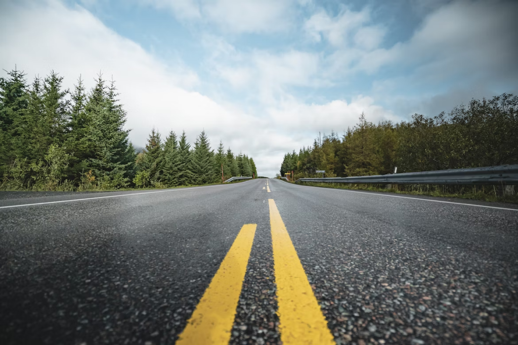 Asphalt road with yellow lines in the center, surrounded by green pine trees and a cloudy blue sky