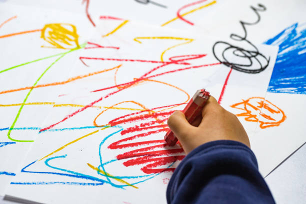 A child's hand holds a red crayon and scribbles on a white sheet of paper. Other colorful scribbles are in the background.
