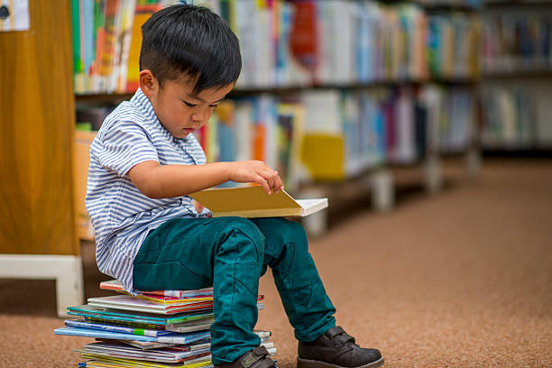 A little boy sits on a pile of books in the library and is looking down at a book. 