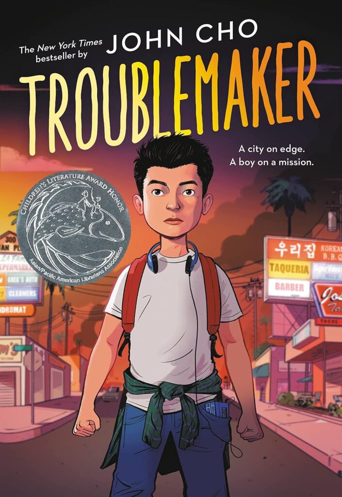 Book cover for "Troublemaker" by John Cho. On the cover a boy is standing wearing a backpack and headphones.