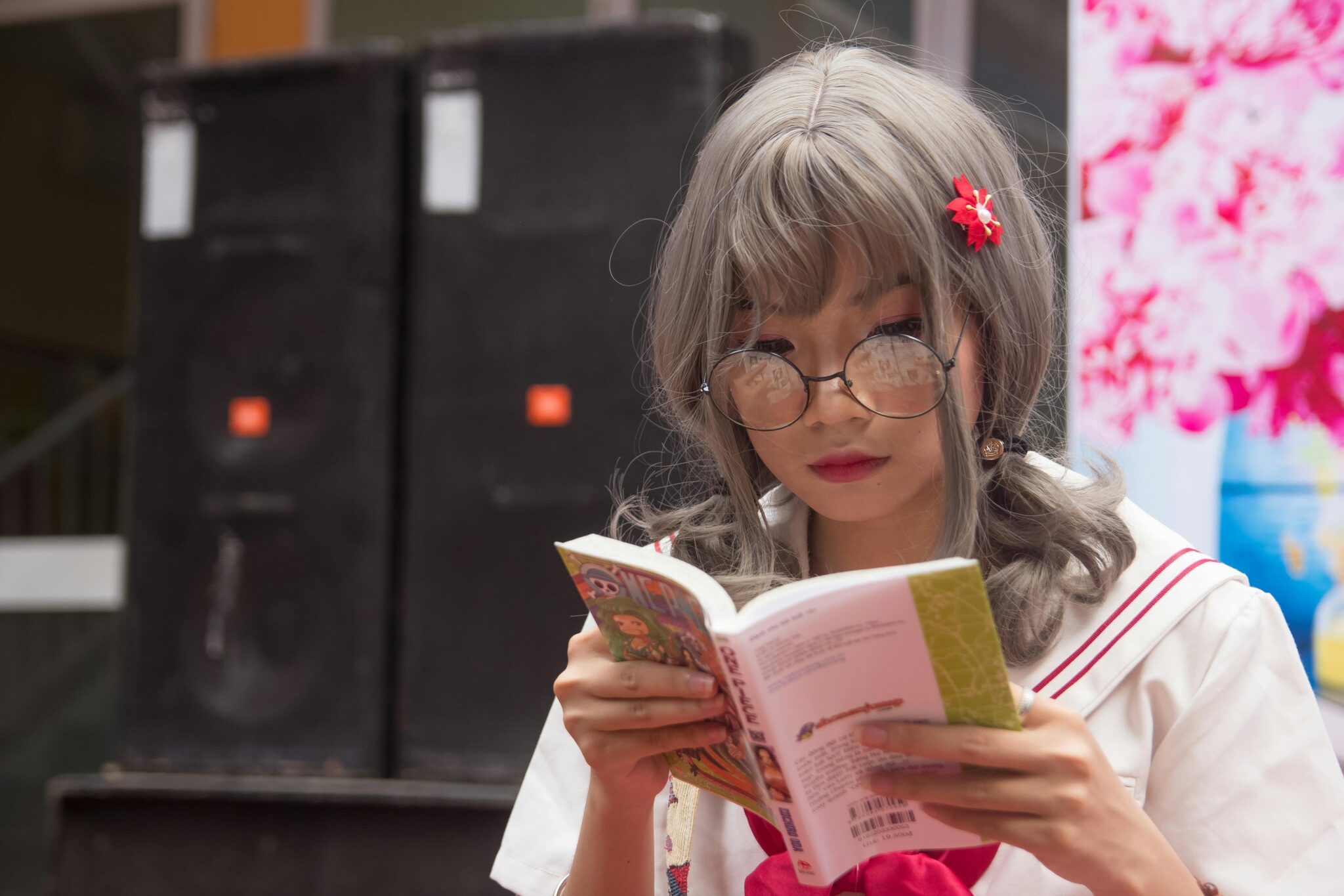 An Asian young adult with medium length gray hair and glasses, dressed in a sailor-style outfit, is intently reading a volume of the One Piece manga. They have a red flower hair accessory. The background is out of focus.