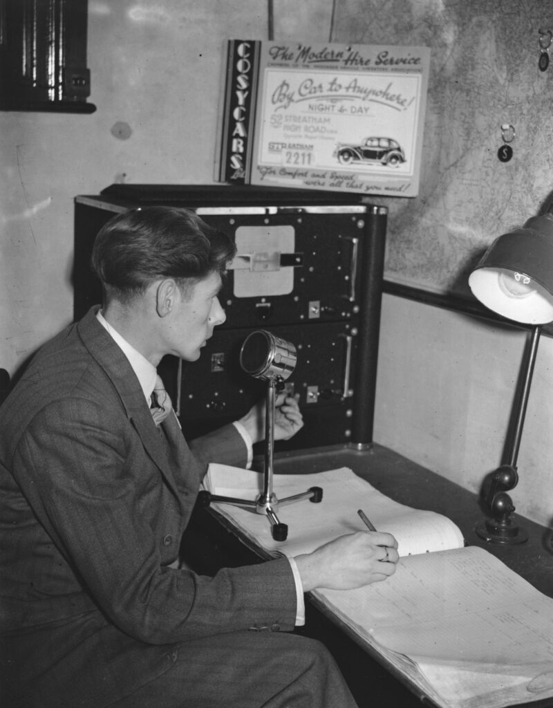 Black and white image of man sitting at desk speaking into an old fashioned microphone.