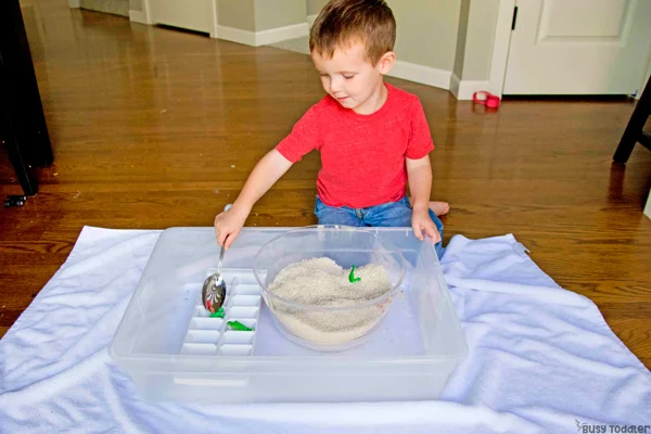 An image of a little boy playing with a spoon, bowl, rice, and dinosaurs on top of a carpet.
