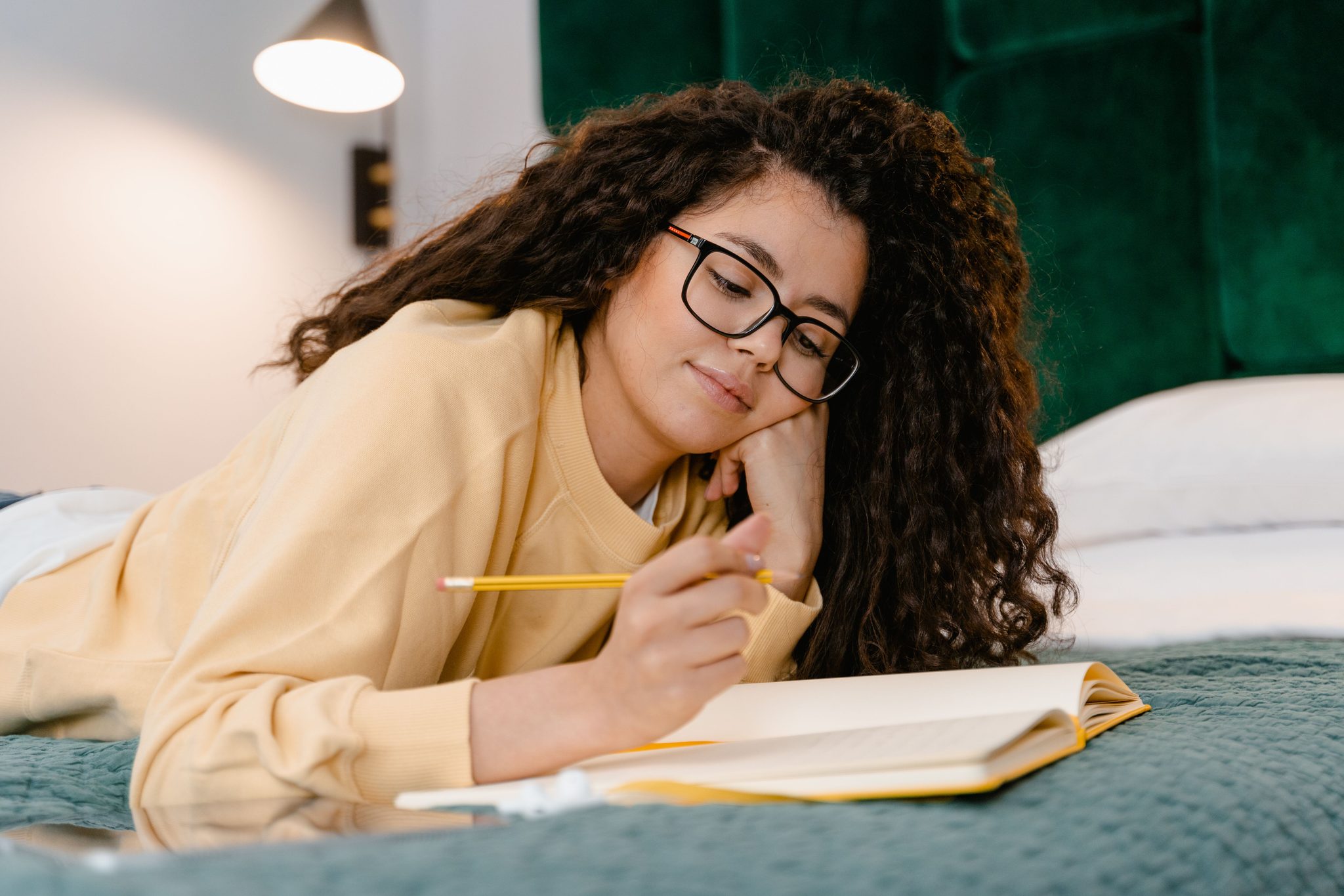 Young woman with curly hair and glasses studying in bed, writing in a notebook. Casual and focused ambiance.